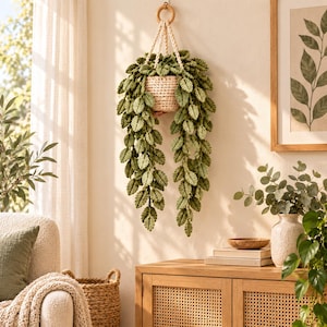 Puede incluir: Una planta colgante de ganchillo con una maceta de color crema y largas enredaderas con hojas verdes texturizadas. La planta está suspendida por un cordón de macramé de color crema sujeto a un anillo de madera circular para colgar en la pared.