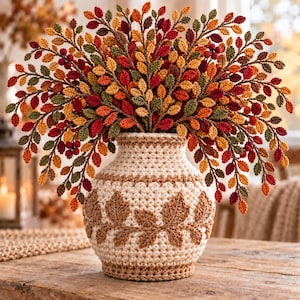 May include: A crochet vase in cream and tan with a textured leaf pattern, filled with a large arrangement of crocheted autumn-colored branches featuring small leaves and berries in shades of red, orange, yellow, brown, and dark green.