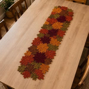 May include: A rectangular table runner composed of interconnected crocheted leaf motifs in shades of orange, yellow, olive green, brown, and deep burgundy. The intricate lace-like pattern features various maple leaf shapes joined together in a long strip.