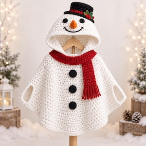 May include: A white crocheted poncho featuring a hood designed as a snowman face with black eyes, an orange nose, and a smile. The garment includes a red crocheted scarf, three black circular buttons down the front, and a black top hat with holly.