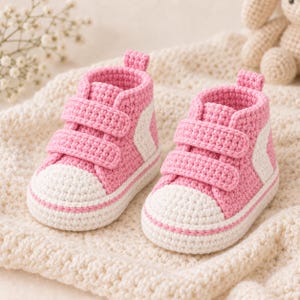 Puede incluir: Un par de zapatillas de ganchillo hechas a mano para bebé en color rosa y blanco. Los zapatos presentan un diseño de caña alta con dos correas de cierre de velcro en la parte delantera, puntera blanca y un detalle lateral blanco, todo en punto.