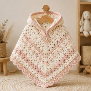 May include: A cream and light pink crocheted hooded poncho featuring a delicate granny square pattern, a scalloped hem, and a single flower-shaped button closure at the neckline. The garment is displayed on a wooden hanger.