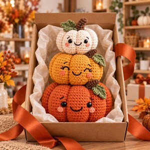 Peut inclure: Une pile de trois citrouilles au crochet dans une bo&icirc;te cadeau en carton doubl&eacute;e de papier de soie blanc. Les citrouilles sont blanches, jaunes et orange, chacune avec un visage souriant, des yeux noirs, une bouche brod&eacute;e et des joues roses.