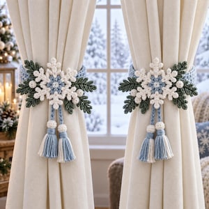May include: A pair of cream-colored curtains held back by two crocheted tiebacks. Each tieback features a large white snowflake center, dark green crocheted leaves, small white berries, and two light blue and white tassels hanging down.