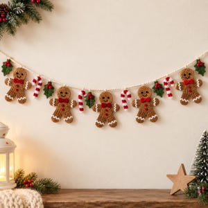 May include: A festive garland featuring five crocheted gingerbread figures with red bow ties and white icing details, interspersed with crocheted candy canes and green holly leaves with red berries, all strung together on a thick white cord.