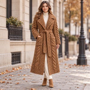 May include: A long, tan, cable-knit crochet coat featuring a wide lapel, long sleeves, two front patch pockets, and a matching tie belt at the waist. The garment is worn over a cream-colored turtleneck sweater and matching cream-colored trousers.