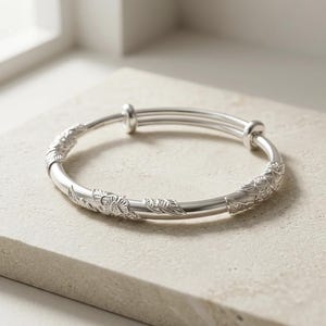 May include: A silver-toned bangle bracelet featuring a smooth, circular band with adjustable overlapping ends. The surface of the metal is decorated with embossed floral and leaf patterns at intervals, creating a textured, metallic finish.