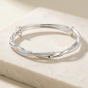 May include: A silver-toned bangle bracelet featuring a multi-strand design with two smooth, polished bands intertwined with a single textured, glittery band. The circular bracelet has a continuous, overlapping structure with rounded ends.