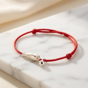 Peut inclure: Un bracelet en cordon rouge doté d'une breloque en forme de poisson argenté lisse et d'une petite perle ronde argentée. Le bracelet est fabriqué à partir d'un fin cordon rouge tressé avec des nœuds réglables, offrant un design épuré et simple.