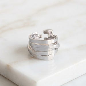 Puede incluir: Un anillo de tono plateado con un diseño envolvente de múltiples capas. La banda consta de varias tiras de metal planas, lisas y superpuestas que crean una apariencia ancha y apilada con un espacio abierto en la parte superior.