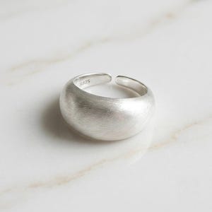 May include: A silver-colored open ring with a wide, rounded band and a brushed, matte texture. The interior of the band is clearly stamped with the text S925, indicating the metal composition. The ring features a gap at the back for adjustment.