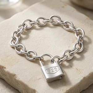 May include: A silver chain link bracelet featuring a polished padlock charm. The padlock is engraved with the number 925, indicating sterling silver composition. The bracelet consists of uniform circular links connected in a continuous loop.