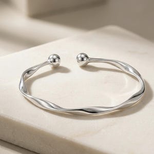 Può includere: Un bracciale aperto color argento lucido caratterizzato da un design a fascia metallica ritorta continua. Il bracciale termina con due terminali sferici lisci rivolti l'uno verso l'altro, creando un'apertura nella struttura circolare.