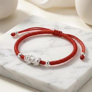 Può includere: Un bracciale in corda intrecciata rossa con un ciondolo centrale a forma di creatura mitica color argento, affiancato da due piccole perline color argento. Presenta una chiusura a nodo scorrevole regolabile con due cordini e perline finali.