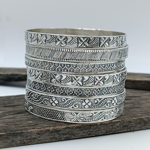 May include: A stack of six silver-toned metal bangles, each featuring a distinct embossed pattern. The designs include repeating floral motifs, geometric cross-hatch textures, and intricate scrollwork, all finished with a polished metallic sheen.
