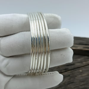 Puede incluir: Un conjunto de siete brazaletes finos y pulidos de tono plateado apilados juntos. Las pulseras presentan un perfil liso y redondeado con un acabado metálico reflectante, creando una apariencia uniforme en todo el conjunto de joyería.