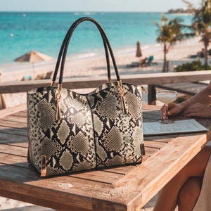 May include: A tote bag featuring a grey and black snakeskin pattern with a textured finish. The bag includes two dark brown braided handles attached with gold-tone metal hardware, and gold-tone metal corner accents at the base of the bag.