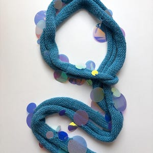 May include: A teal knitted scarf featuring numerous circular, iridescent sequins attached along the length of the fabric. The scarf has a textured, ribbed knit construction and the sequins reflect various colours including blue, purple, and silver.