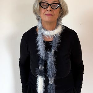 May include: A long, thin scarf featuring a mix of fuzzy, textured yarn in shades of white, light gray, and dark charcoal gray. The scarf is accented with small, reflective silver sequins interspersed throughout the length of the soft, feathery material.