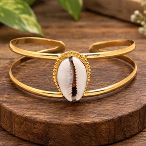 May include: A gold-toned open cuff bracelet featuring a central white cowrie shell. The shell is framed by a beaded gold-colored border, and the bracelet consists of two parallel bands that meet at the sides to form a single open-ended structure.