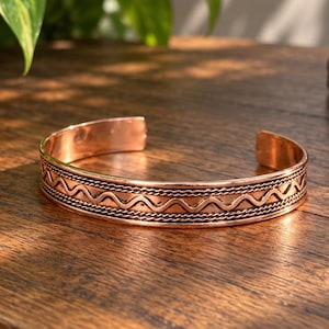 May include: A copper cuff bracelet featuring a central horizontal wavy line pattern flanked by two thin, textured rope-like borders. The metal has a polished, warm metallic finish with dark oxidation highlighting the intricate engraved details.