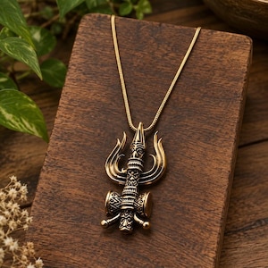 Puede incluir: Un collar con colgante de metal en tono dorado que presenta un diseño en forma de tridente con intrincados patrones grabados y una pequeña estructura similar a un tambor en la base. El colgante cuelga de una fina cadena de serpiente a juego.