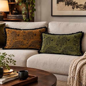 May include: Two rectangular throw pillows with black fringe trim. One pillow is mustard yellow and the other is olive green, both featuring a repeating black leopard pattern. The pillows are positioned on a light-colored sofa.