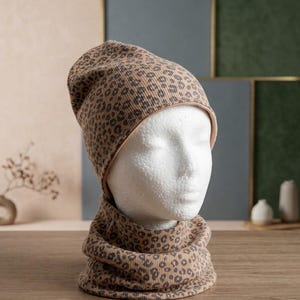 Könnte beinhalten: Ein Set aus einer gerippten Strickmütze und einem passenden Schlauchschal in Beige mit einem dunkelbraunen Leopardenmuster. Das Material weist durchgehend eine vertikale Rippenstruktur auf, präsentiert auf einem weißen Kopfmodell.