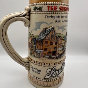 May include: A beige ceramic beer stein featuring a relief scene of a German village with half-timbered houses. The text reads THE STROH, During the late 18th, Kirn, Germany, and Heritage Series II. It has a curved handle and a tan rim.