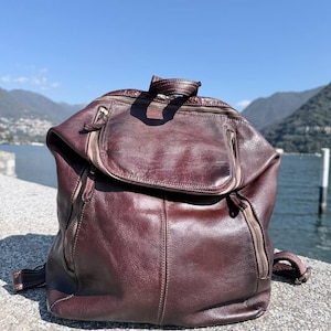 Könnte beinhalten: Ein dunkelbrauner Lederrucksack mit einer großen oberen Klappe mit Reißverschluss, zwei vertikalen Reißverschlusstaschen auf der Vorderseite, einem Tragegriff und verstellbaren Schultergurten mit Metallbeschlägen und sichtbaren Nähten.