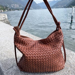 Può includere: Una borsa a mano marrone di medie dimensioni caratterizzata da un intricato motivo in pelle intrecciata. La borsa presenta una struttura morbida e flessibile con due sottili tracolle in pelle marrone abbinate fissate sulla parte superiore.