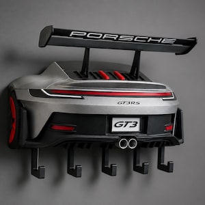 Puede incluir: Un llavero de pared plateado con forma de la parte trasera de un coche deportivo Porsche GT3 RS. Cuenta con un gran alerón trasero negro con el texto PORSCHE, detalles en rojo, tubos de escape dobles y cinco ganchos negros en el borde inferior.
