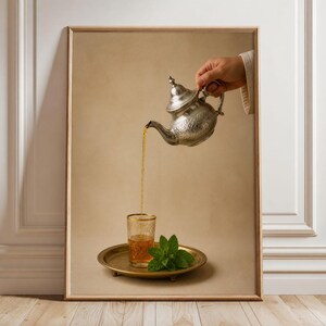 Puede incluir: Una impresión de arte mural enmarcada que muestra una tetera de plata vertiendo té de color ámbar en un vaso sobre una bandeja de latón. Junto al vaso hay una ramita de hojas de menta verde fresca. La escena se sitúa sobre un fondo beige neutro.