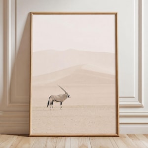 Può includere: Una stampa artistica da parete rettangolare in una cornice di legno chiaro che raffigura una fotografia di un orice in piedi in un vasto e pallido paesaggio desertico con morbide dune di sabbia sullo sfondo sotto un cielo fosco.