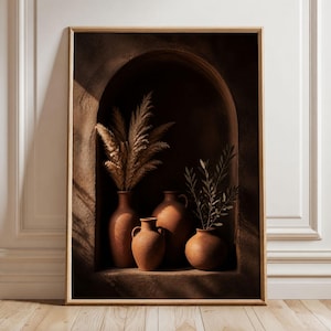May include: A framed wall art print featuring an arched alcove containing four terracotta-coloured clay vases. Two vases hold dried pampas grass plumes, while one holds a small green leafy branch. The scene is lit with warm, directional sunlight.