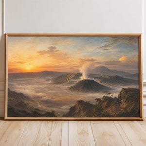 May include: A rectangular landscape painting in a light wood frame depicting a volcanic mountain range at sunrise. The scene features a central smoking volcano surrounded by mist and rugged terrain under a golden, orange, and blue sky.