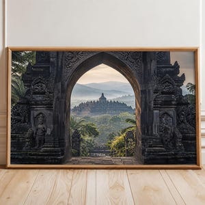 May include: A rectangular wall art print in a light wood frame featuring a dark stone archway framing a view of the Borobudur temple complex. The scene shows the ancient structure surrounded by lush green trees under a soft, hazy morning sky.