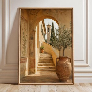 Op de afbeelding: Een ingelijste kunstdruk met een mediterrane stenen boog die naar een trap leidt, een hoge terracotta urn met een olijfboom en een verre klokkentoren. De scène bevat warme beige en okerkleurige tinten met gedetailleerde architecturale texturen.