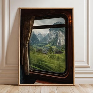Può includere: Una stampa artistica da parete incorniciata che mostra una vista dal finestrino di un treno. La scena ritrae una valle verdeggiante con piccole case sotto imponenti montagne innevate. Una tenda marrone pende a sinistra del finestrino.