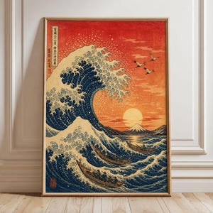 May include: A framed vertical art print featuring a large dark blue ocean wave, three wooden boats, a bright sun, and Mount Fuji against a vibrant orange sky. Japanese text is visible in the upper left corner of the print, which is set in a light wood frame.