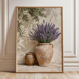 Könnte beinhalten: Ein gerahmter Kunstdruck mit einer großen Terrakotta-Vase voller violetter Lavendelzweige neben einem kleineren, abgerundeten Tontopf. Die Szene ist vor einer strukturierten beigen Wand platziert, auf die Schatten von Olivenbaumzweigen fallen.