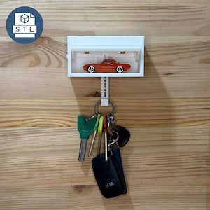 May include: A white rectangular wall-mounted key holder featuring a small orange toy sports car displayed inside the top compartment. A vertical extension hangs below the main unit, supporting a metal ring with several keys and a black car key fob.
