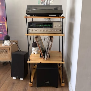 May include: A four-tier audio equipment rack constructed from light-toned wood shelves and black metal support rods. The unit holds a black turntable on the top shelf, a silver stereo receiver with green display lights, and a vinyl record storage rack.