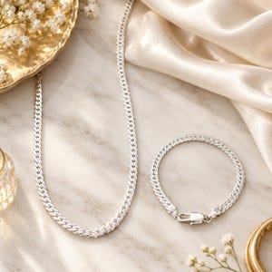 Può includere: Una collana a catena gourmette in argento e un bracciale a catena gourmette in argento abbinato. Entrambi i pezzi presentano un design a maglie piatte intrecciate con una finitura lucida e riflettente. Il bracciale include una chiusura a moschettone.