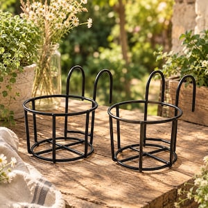 Può includere: Due supporti per vasi da fiori in metallo nero con base circolare e un gancio verticale per appenderli. Ogni supporto presenta una semplice struttura in filo metallico progettata per sostenere un contenitore, con un design pulito.