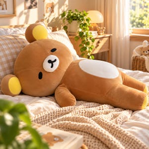Pode incluir: Um grande e macio urso de pelúcia castanho com uma mancha oval branca na barriga, um focinho branco, olhos pretos e o interior das orelhas amarelo. O urso está deitado horizontalmente numa cama, com uma cabeça arredondada e pequenas orelhas.