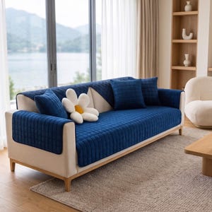 Puede incluir: Un sofá de color crema cubierto con un protector de tela acolchada azul intenso. El sofá cuenta con cojines cuadrados azules a juego y un cojín decorativo blanco en forma de flor con el centro amarillo sobre los asientos.