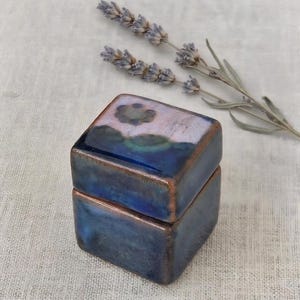 Puede incluir: Una pequeña caja cuadrada de cerámica con un esmalte brillante en azul profundo y morado. La tapa presenta un motivo floral circular oscuro y estilizado sobre un fondo lavanda más claro. Los bordes tienen un acabado metálico color cobre.