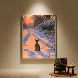 Puede incluir: Una impresión de arte mural rectangular que muestra una liebre marrón sentada en un paisaje cubierto de nieve durante un vibrante atardecer naranja. La escena incluye un camino con huellas, arbustos nevados con bayas rojas y árboles.