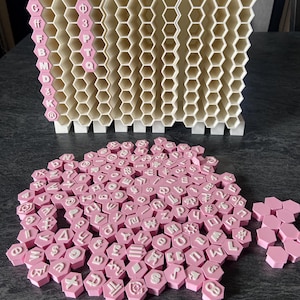 May include: A white 3D-printed honeycomb storage rack with hexagonal compartments and a collection of pink hexagonal tiles featuring white embossed letters, numbers, and symbols scattered in front of the rack.